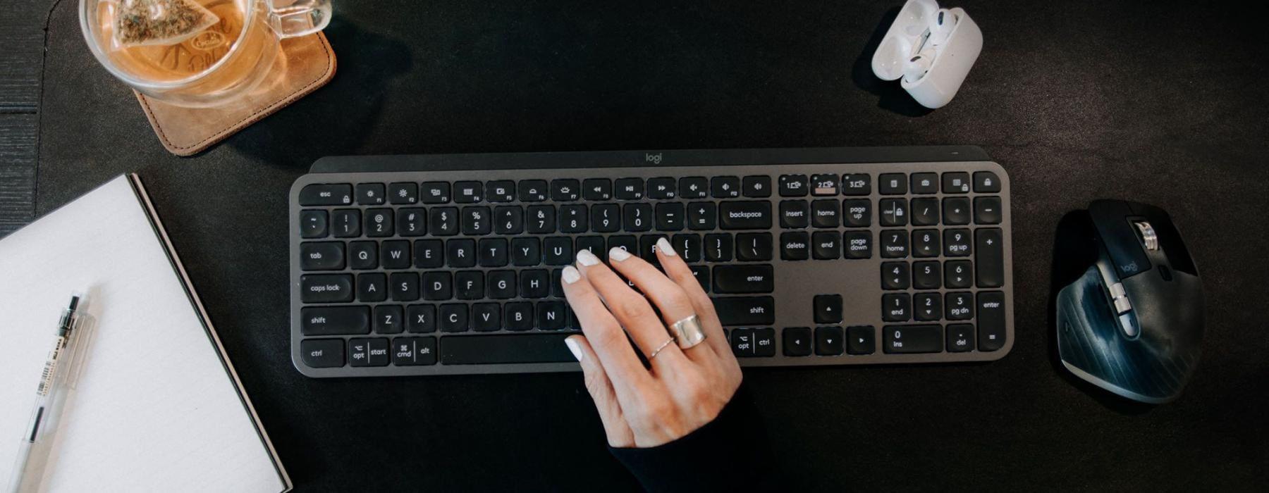 woman's hand on a keyboard surrounded by office items and a cup of tea on a coaster